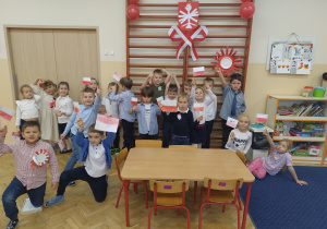 Dzieci stoją na tle symboli narodowych z kotylionami i flagami