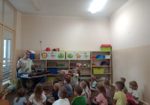 Grupa siedzi na dywanie po turecku i słucha pana grającego na keybordzie