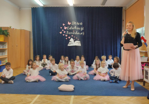 Pani stoi z mikrofonem, dzieci siedzą