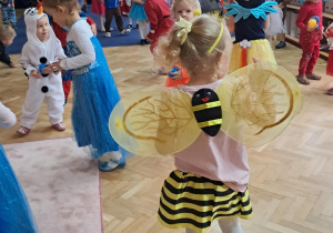 Dziewczynka stojąca tyłem, przebrana za pszczółkę, w tle dzieci w przebraniach