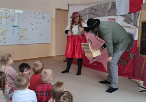 Dwóch aktorów na scenie, jeden z nich trzyma drewnianą szkatułkę.