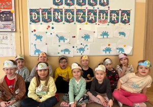 Grupa siedzi pod tablicą z napisem Dzień Dinozaura