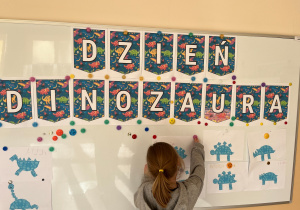 Dziewczynka przyczepia na tablicę wyciętego dinozaura