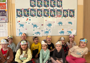Grupa w opaskach z dinozaurami pozuje przed tablicą