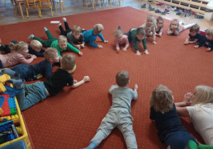 Dzieci wykonują ćwiczenie leżąc na dywanie z rękami przed sobą.