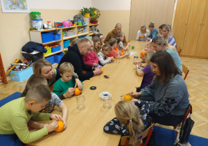 Rodzice siedzą z dziećmi przy stoliku i pomagają im w wykonaniu ozdoby z pomarańczy.