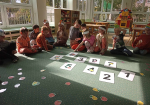 Dzieci siedzą w kole na dywanie i patrzą na karty z liczbami oraz rozsypankę z owocami.