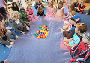 Dzieci siedzą w kole i trzymają chustę na której znajdują się kolorowe piłeczki.