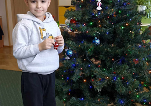 Chłopiec stoi przy choince z Mikołajem w dłoni.